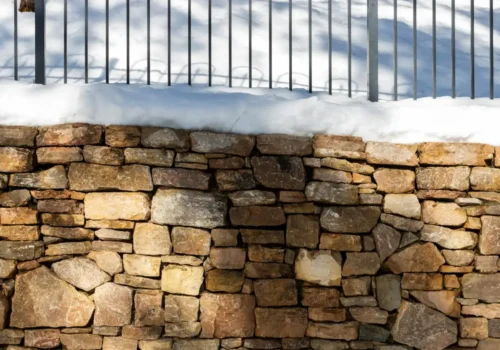 A stone wall covered in snow next to a fence, block foundation in Greenville NC