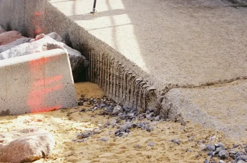Cracked concrete slab with exposed steel rebar above sand and dark pebbles, concrete footing